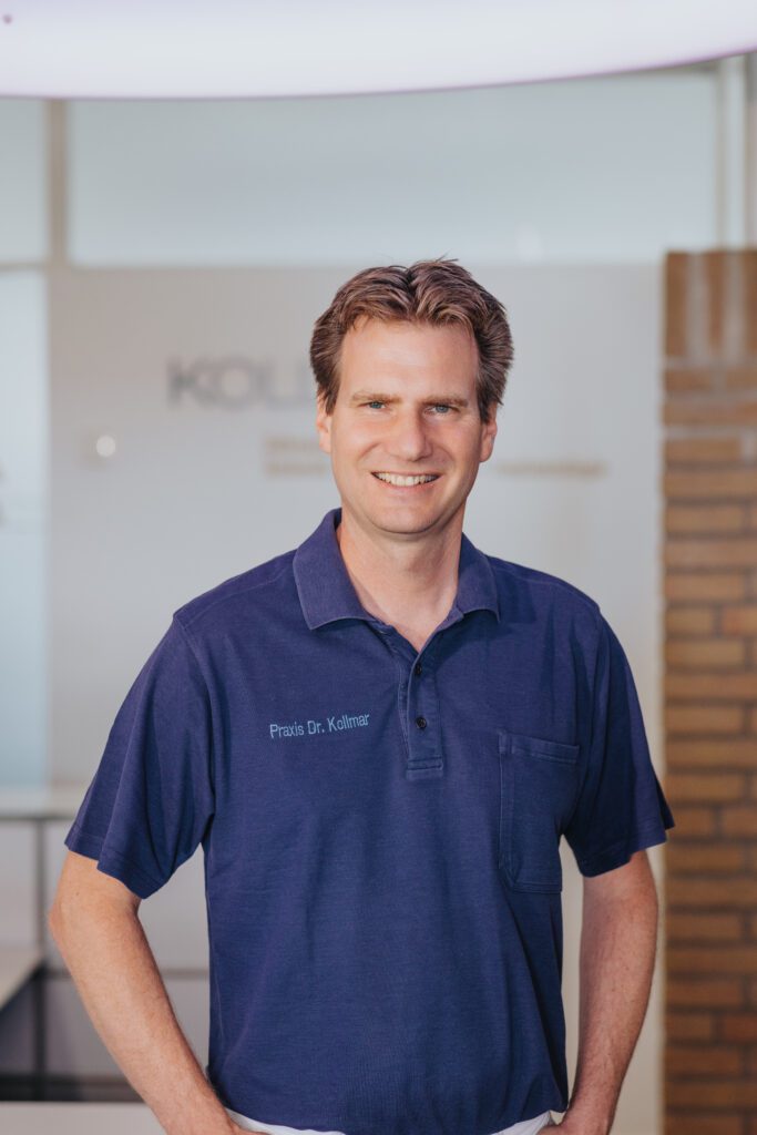Porträt von Zahnarzt Dr. Fritz Kollmar vor dem Logo seiner Zahnarztpraxis in Kassel – freundlicher Empfang für Patienten.